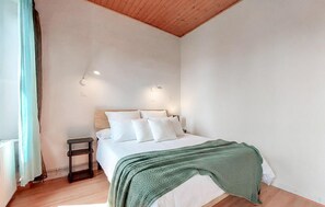 A bedroom with a wooden ceiling, a bed with white bedding, two wall-mounted lights, and a green throw blanket. - Charmante maisonnette (La Rochelle)