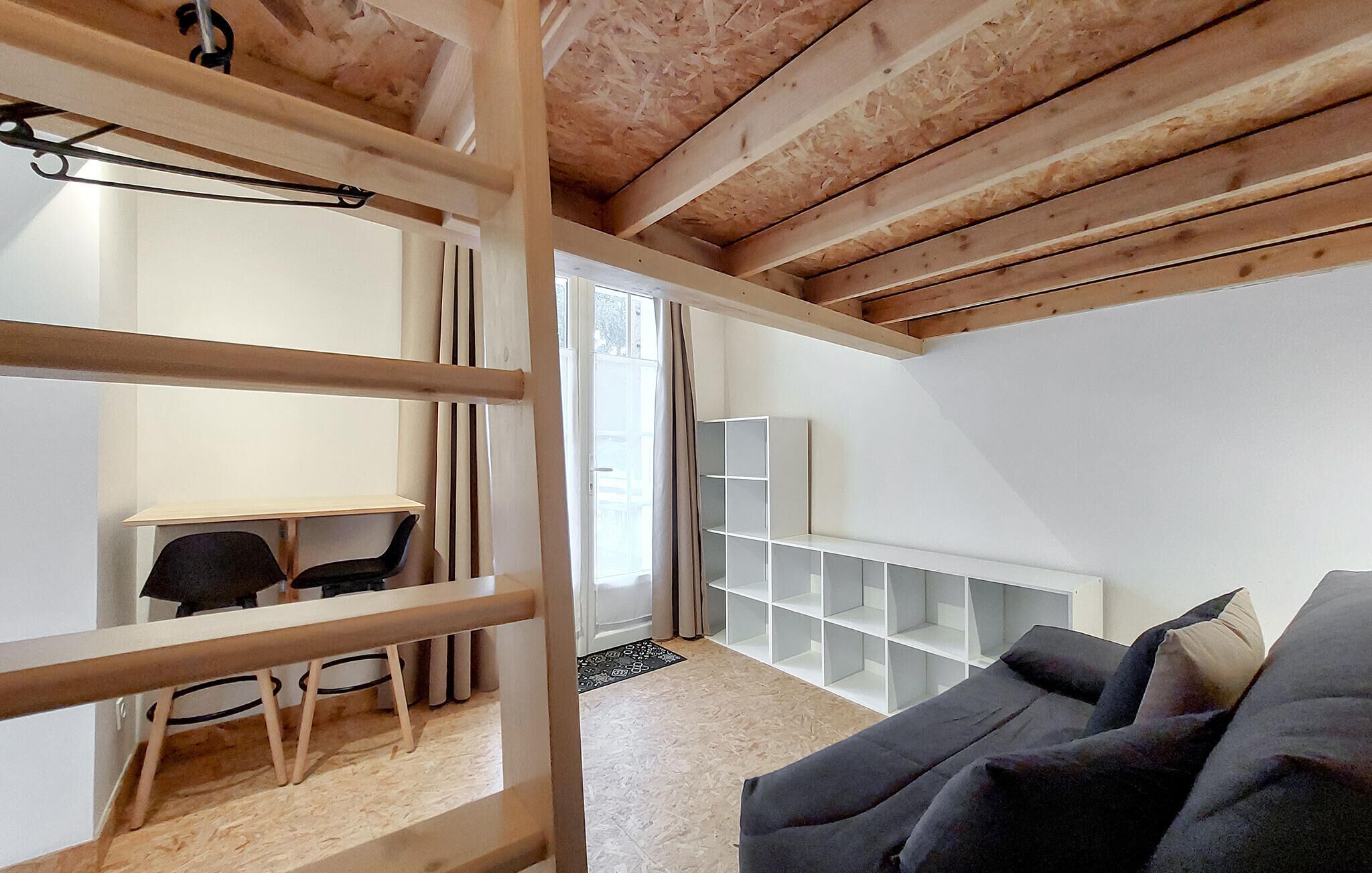 Een kamer met een houten plafond, een zwarte bank, een wit schap en een bureau met een stoel.