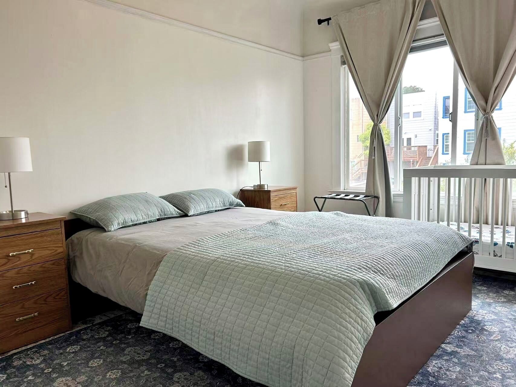 A bedroom with a bed, bedside tables, a lamp, a window with curtains, and a view of buildings outside.