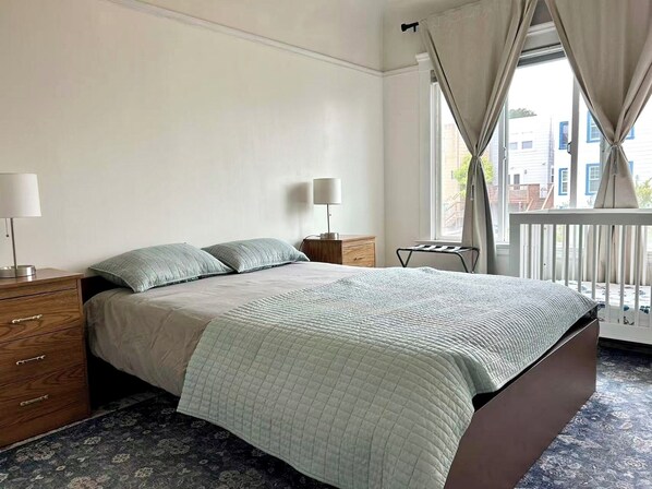 A bedroom with a bed, bedside tables, a lamp, a window with curtains, and a view of buildings outside.