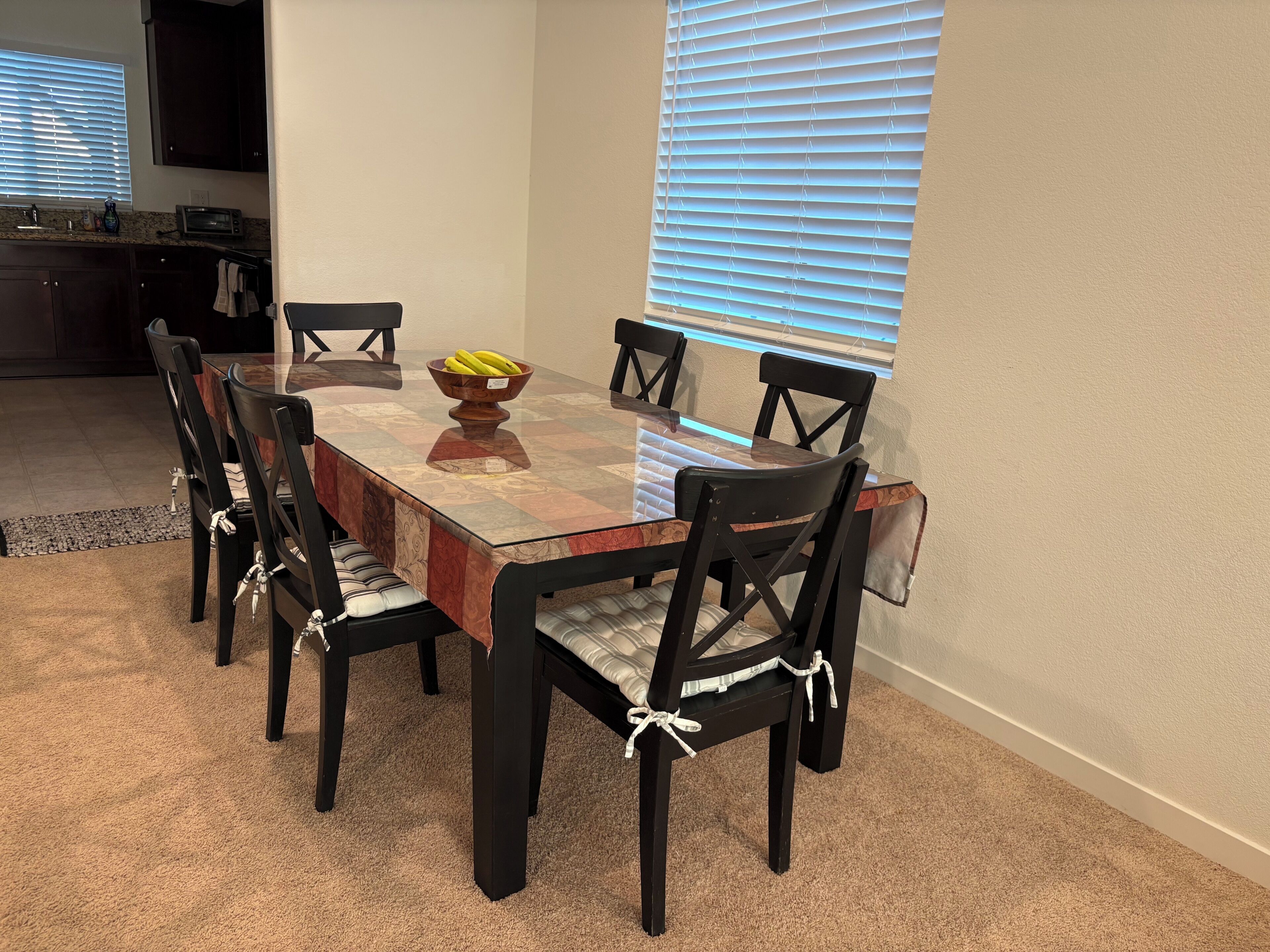 A dining area with a glass-top table, six chairs, a bowl of fruit, and a window with blinds.