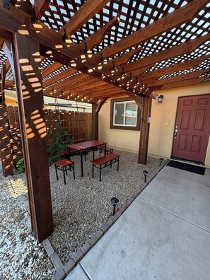 An outdoor patio with wooden pergola, tables, benches, and a red door.