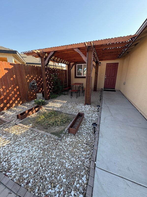 A covered patio area with a wooden pergola, a small table and chairs, and a gravel ground. - Charming 3-bedroom house with Central AC, Pergola, Patio, & Garage in Los Banos. (Los Banos)
