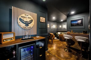 Eine Lounge mit einem Holunderschild mit der Aufschrift 'The Night Owl Lounge', eine Bar mit Hockern, gerahmten Bildern an der Wand und einem Vintage-Radio.
