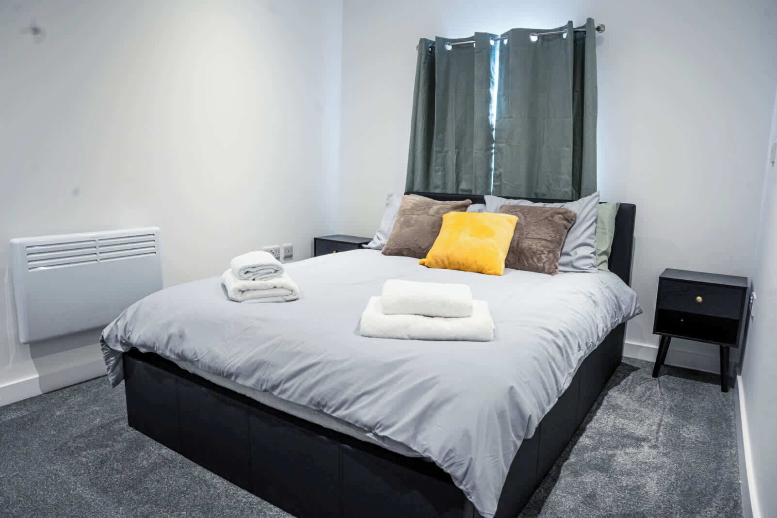 Una cama bien hecha con edredón gris, cabecera negra y una ventana con cortinas verdes.