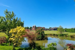 Un paysage rural avec un étang, des arbres et un bâtiment surmonté d’un toit conique.