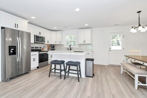 A modern kitchen with white cabinets, stainless steel appliances, a central island with stools, and a dining area with a bench. - The Modern Getaway, 3BR, Workspace + Downtown Kannapolis (Kannapolis)