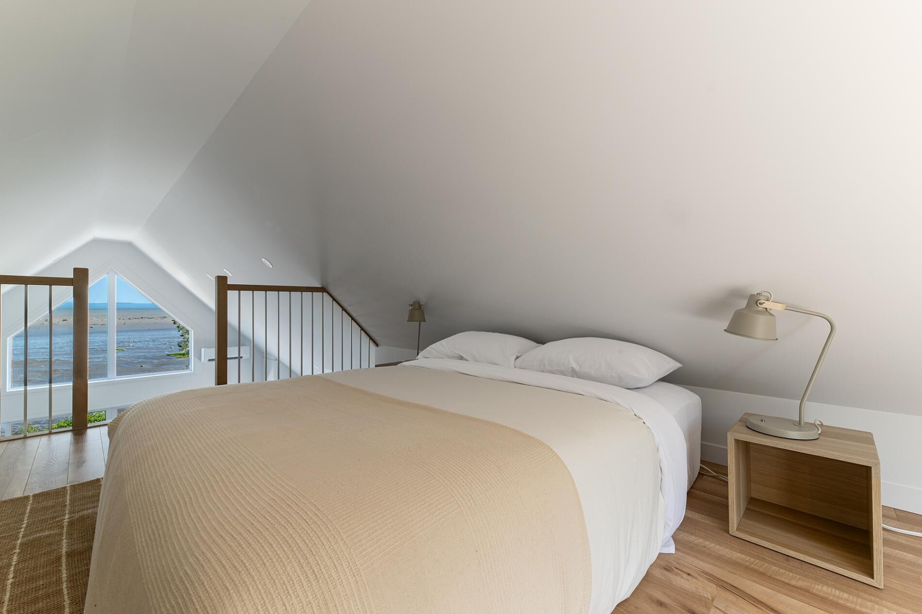 A bedroom with a large bed, a nightstand, a lamp, a window with a view of the beach, and a wooden staircase.