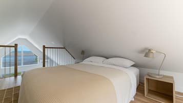A bedroom with a large bed, a nightstand, a lamp, a window with a view of the beach, and a wooden staircase.