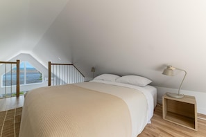 A bedroom with a large bed, a nightstand, a lamp, a window with a view of the beach, and a wooden staircase. - Le Grand Bleu 1265: Waterfront with River Views (Petite-Rivière-Saint-François)