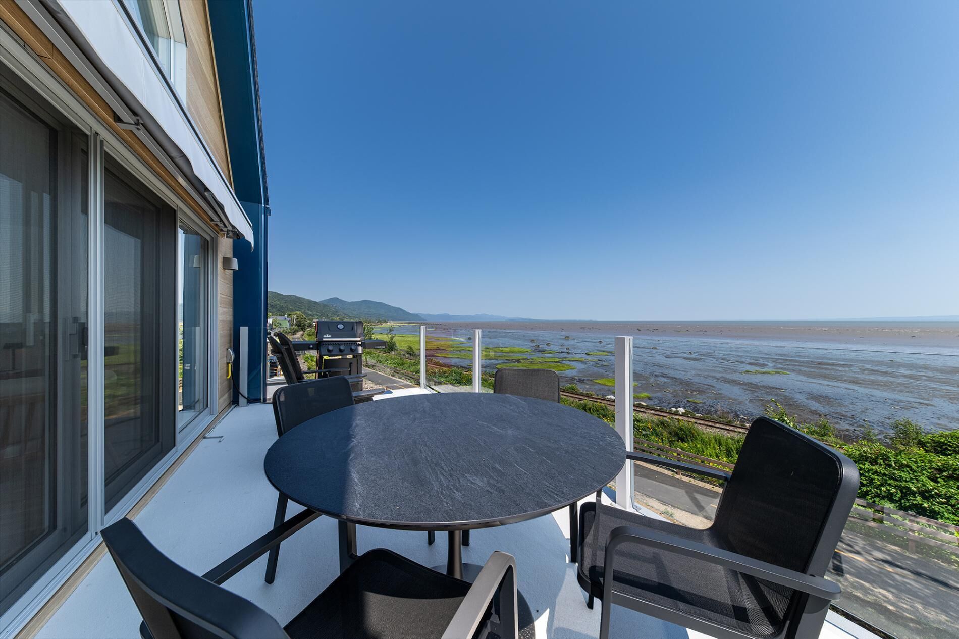 A balcony with a round table and chairs, overlooking a beach and water body.