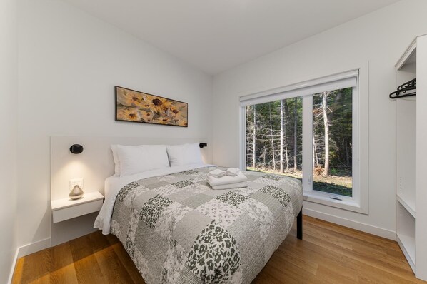 A bedroom with a large bed, a nightstand, a painting, and a window with a forest view.