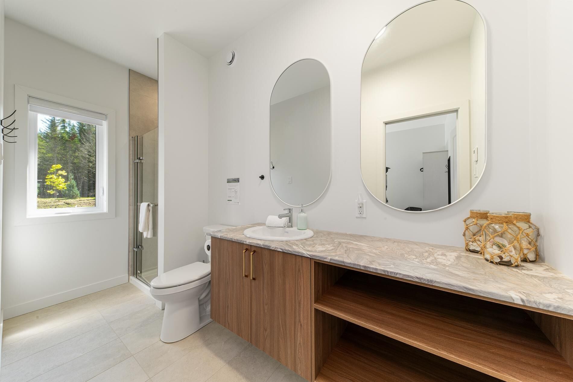 A modern bathroom with a toilet, sink, and vanity. There is a large mirror above the vanity and a window with a view of trees.