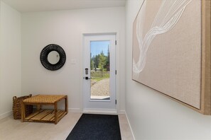 A hallway with a door leading to an outdoor area, a woven bench, a round mirror, and a piece of artwork.