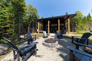 A covered outdoor seating area with Adirondack chairs and a fire pit, surrounded by a forested landscape.