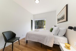A bedroom with a bed, a chair, a bedside table, and a window with a view of trees. - L'Odyssée du Domaine 2: Hot Tub & Firepit (Baie-Saint-Paul)