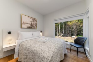 A bedroom with a bed, a chair, a window with a view of trees, and a painting on the wall.