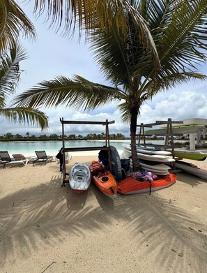 A beach with a row of lounge chairs, a palm tree, and several kayaks and a canoe under a wooden structure. - Tropical Bliss apt, Beach, Golf, Dining,Gated community near downtown Punta Cana (Punta Cana)