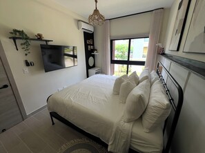 A bedroom with a bed, a TV mounted on the wall, a window with curtains, and a hanging light fixture. - Tropical Bliss apt, Beach, Golf, Dining,Gated community near downtown Punta Cana (Punta Cana)