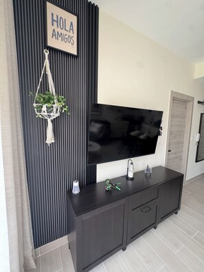 A modern living room with a dark wood console, a flat-screen TV mounted on the wall, a macrame wall hanging, and a 'HOLA AMIGOS' sign. - Tropical Bliss apt, Beach, Golf, Dining,Gated community near downtown Punta Cana (Punta Cana)