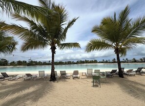A beach with lounge chairs and palm trees, a clear view of the ocean, and a distant shoreline with buildings. - Tropical Bliss apt, Beach, Golf, Dining,Gated community near downtown Punta Cana (Punta Cana)