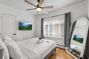 Un dormitorio con cama, ventilador de techo, ventana con persianas, espejo con luces y televisor montado en la pared.