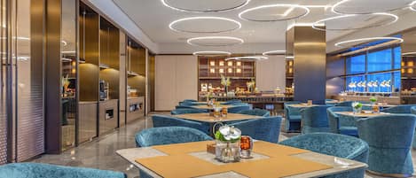 A modern restaurant with blue velvet chairs, round tables, and a bar area with wooden cabinets and decorative plants.