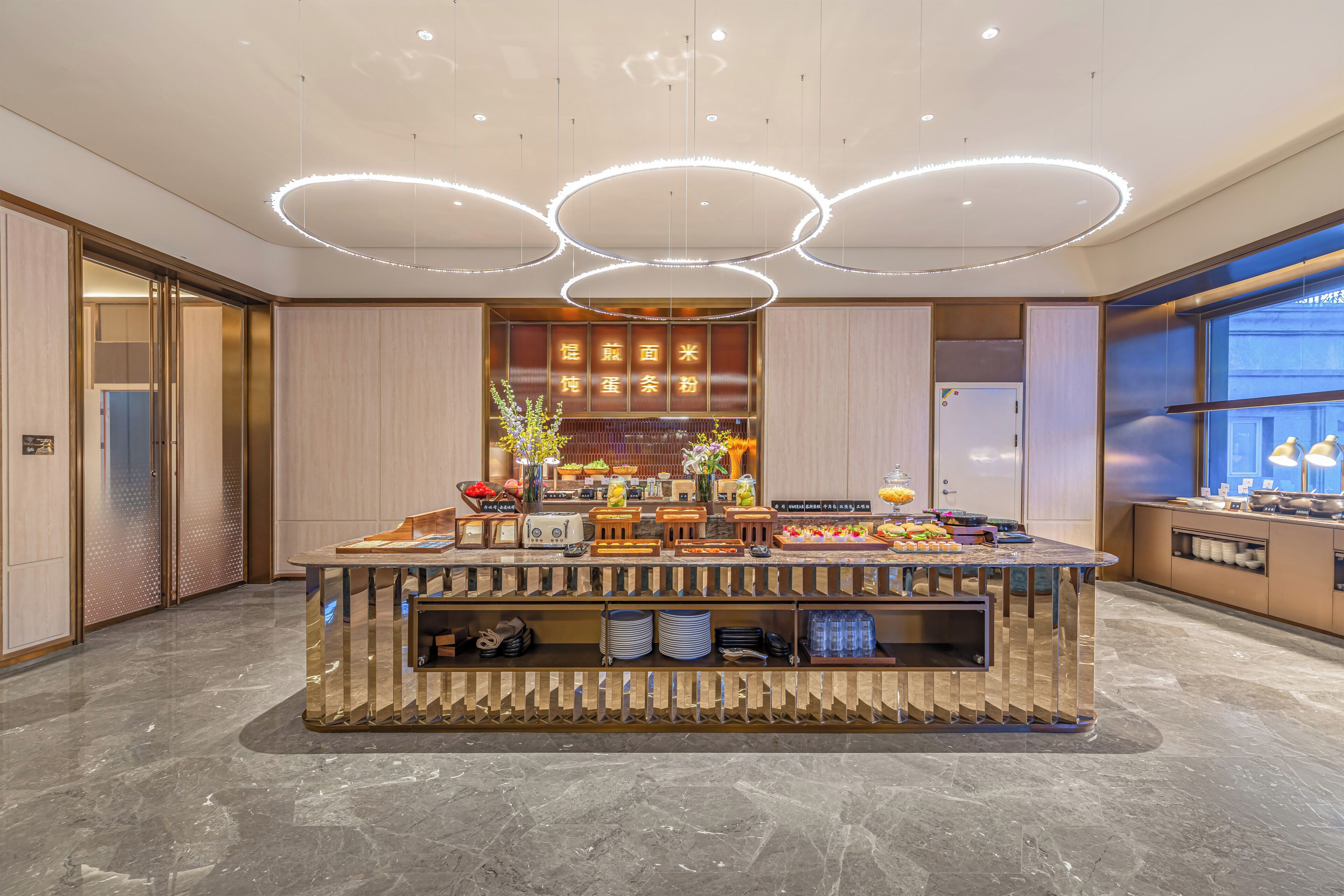 A hotel lobby with a central buffet area, a reception desk, and a large window providing a view of the cityscape.