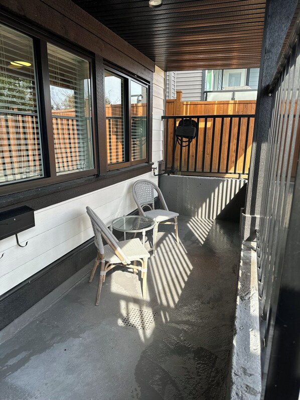 A covered outdoor seating area with a table and two chairs, a wooden fence in the background, and a window with blinds.