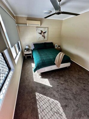 A bedroom with a bed, a nightstand, a wall-mounted air conditioner, a ceiling fan, and a window with a view.