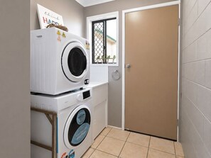 A laundry room with two stacked washing machines, a window with a view of a building, and a closed door.
