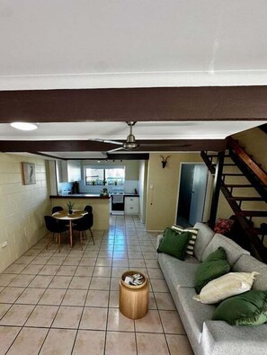 A living room with a sofa, a dining area with chairs, a kitchen in the background, and a staircase.