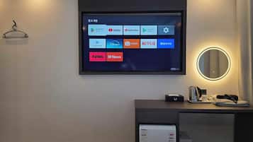A hotel room with a mounted TV displaying various streaming apps, a bedside table with a kettle, a cup, and a remote control, and a wall-mounted mirror with a light.