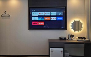 Ein Hotelzimmer mit an der Wand montiertem Fernseher, der verschiedene Streaming-Apps anzeigt, einem Nachttisch mit Wasserkocher, Tasse und Fernbedienung sowie einem beleuchteten Wandspiegel.