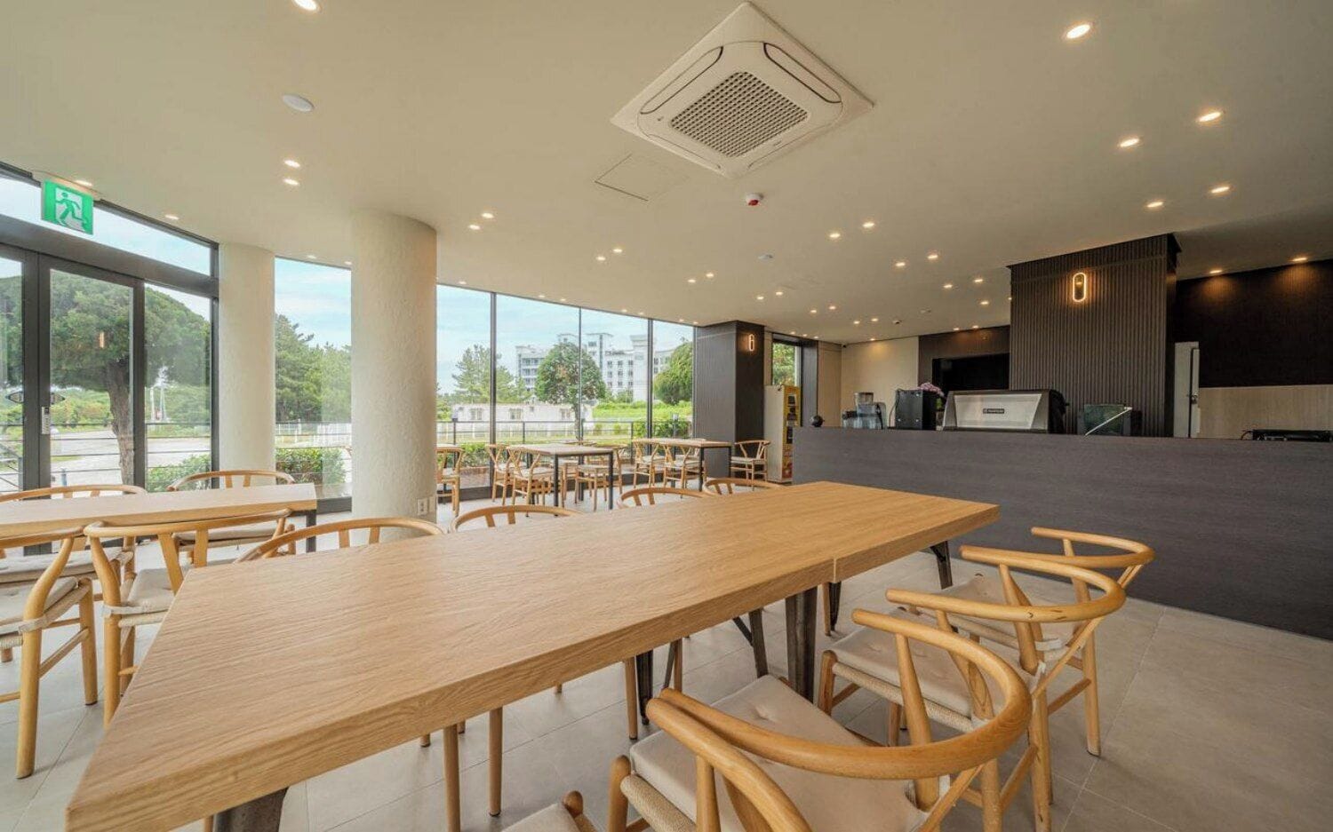 A spacious indoor area with multiple tables and chairs, large windows offering a view of the outdoors, and a reception desk in the background.