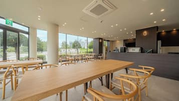 A spacious indoor area with multiple tables and chairs, large windows offering a view of the outdoors, and a reception desk in the background.