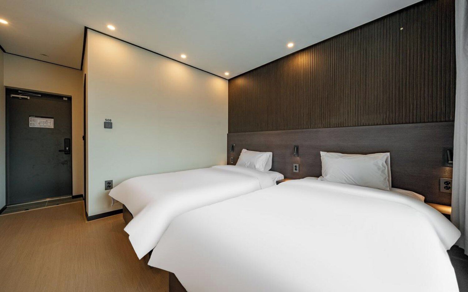 A hotel room with two beds, a wooden headboard, a door with a sign, and a light switch on the wall.