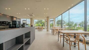 A modern cafe with a long counter, wooden tables, and large windows offering a view of greenery.