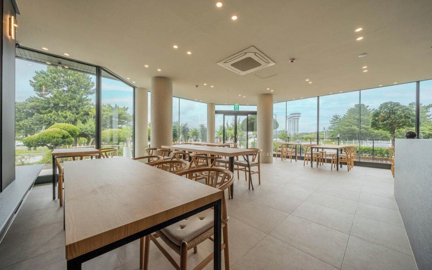 A spacious indoor area with multiple wooden tables and chairs, large windows offering a view of greenery outside.
