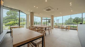 A spacious indoor area with multiple wooden tables and chairs, large windows offering a view of greenery outside.