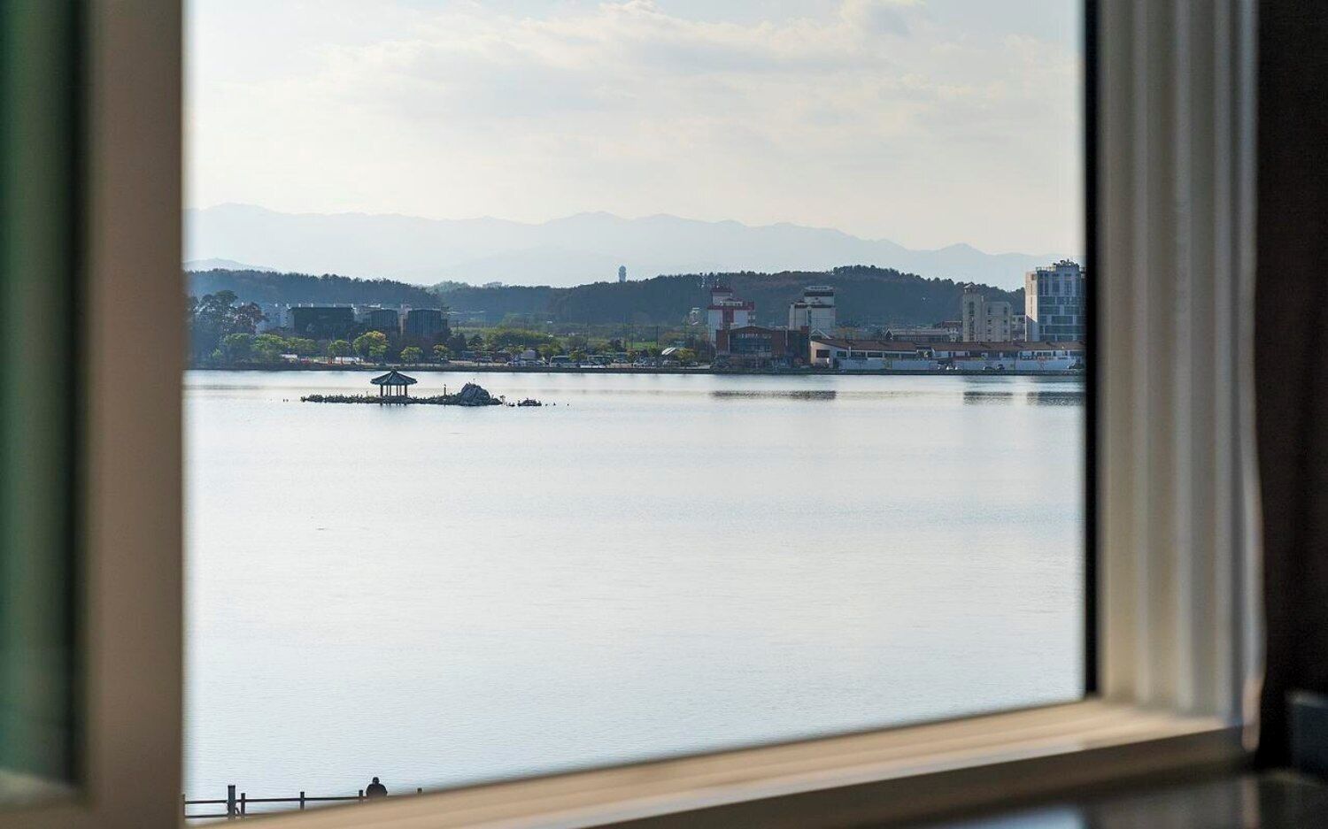 A view of a calm body of water with a small island and a gazebo, buildings in the distance, and mountains in the background.