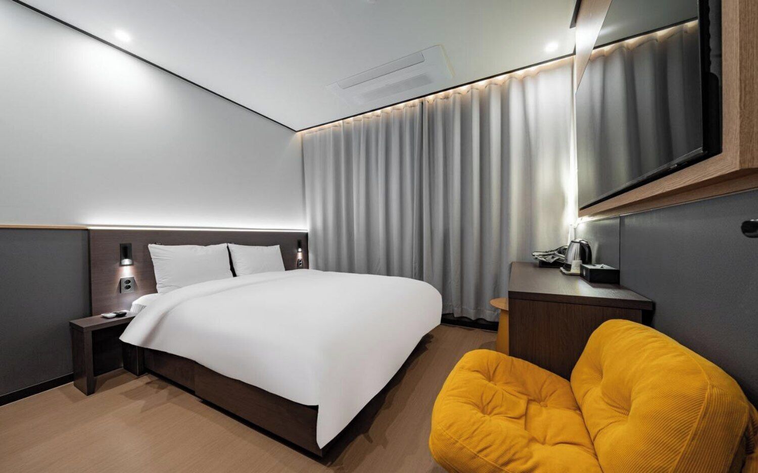 A modern hotel room with a large bed, a bedside table, a chair, a desk, and a mirror.