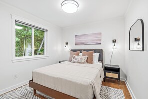 Een slaapkamer met een bed, nachtkastjes, een raam met uitzicht op bomen, een wandlamp en een schilderij.
