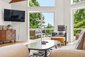 Een moderne woonkamer met een flatscreen televisie, een salontafel met boeken, een bank, een fauteuil en uitzicht op groen door grote ramen.