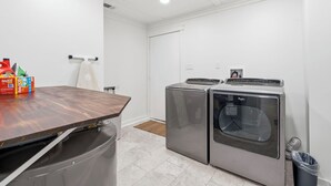 A laundry room with two front-loading washers and dryers, a wooden table, and a trash can.