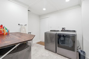 A laundry room with two front-loading washers and dryers, a wooden table, and a trash can.