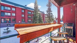 Um deck de madeira com um banco e banquetas, com vista para um pátio coberto de neve e edifícios ao fundo.