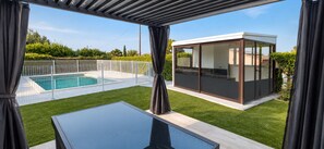 Et moderne poolområde udendørs med en glasinddækket konstruktion, en klar himmel og velplejet græsplæne.