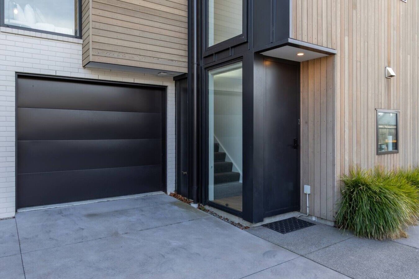 A modern two-story house with a dark garage door, a glass entrance, and a small outdoor area with a planter.