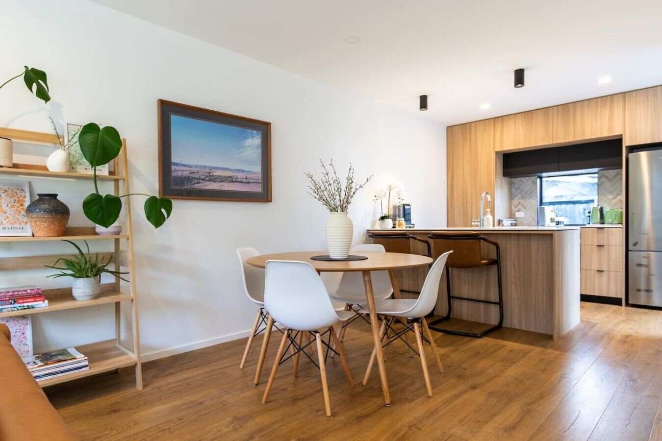 Una cocina moderna con armarios de madera, un refrigerador de acero inoxidable y un comedor con una mesa redonda y sillas blancas.